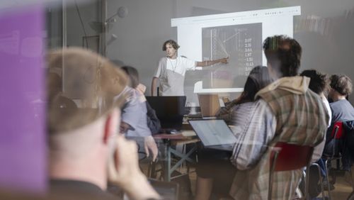 workshop situation. A person explains something pointing finger towards schematic representation. other people are looking at the person, giving their back to the camera
